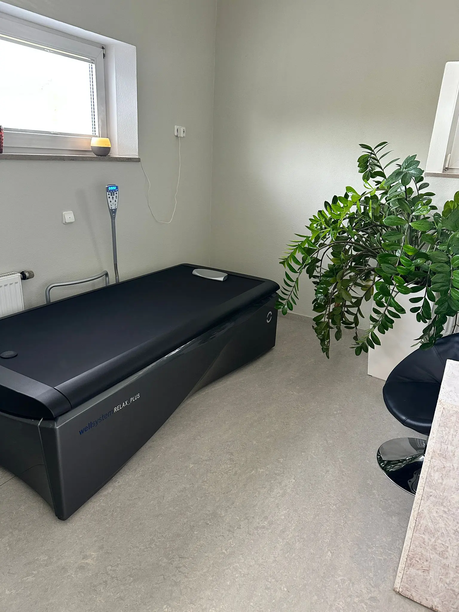 Ein elegantes schwarzes Hydromassagebett steht in einem minimalistischen Raum mit hellen Wänden, einer großen Blattpflanze, einem modernen schwarzen Stuhl und einem Fenster, das Tageslicht hereinlässt.