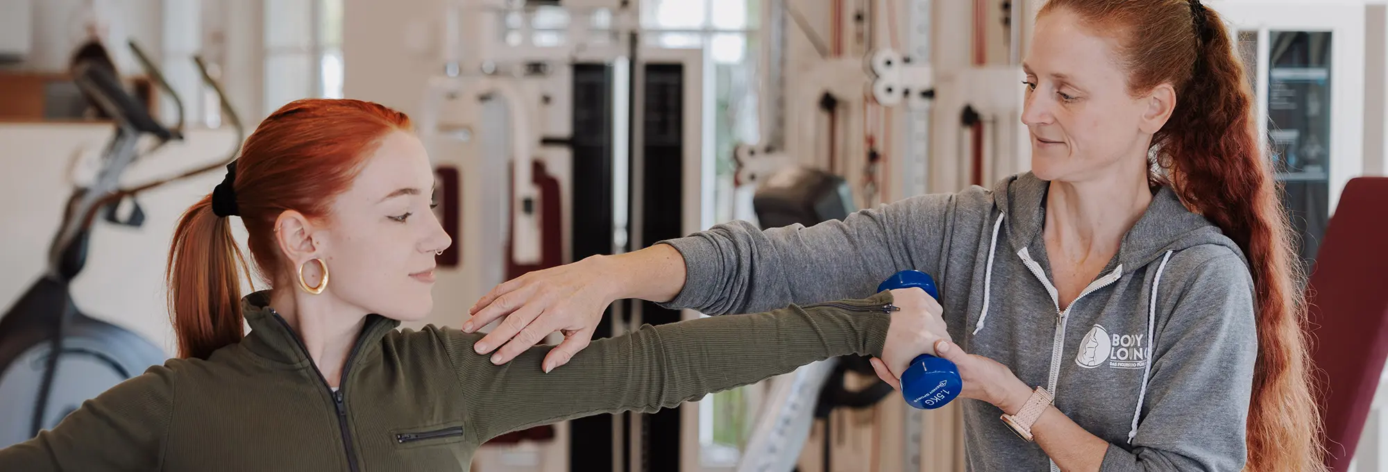 Eine Frau in Sportkleidung hilft einer anderen Frau beim Heben einer blauen Hantel und führt ihren Arm während eines Trainings in einem Fitnessstudio.